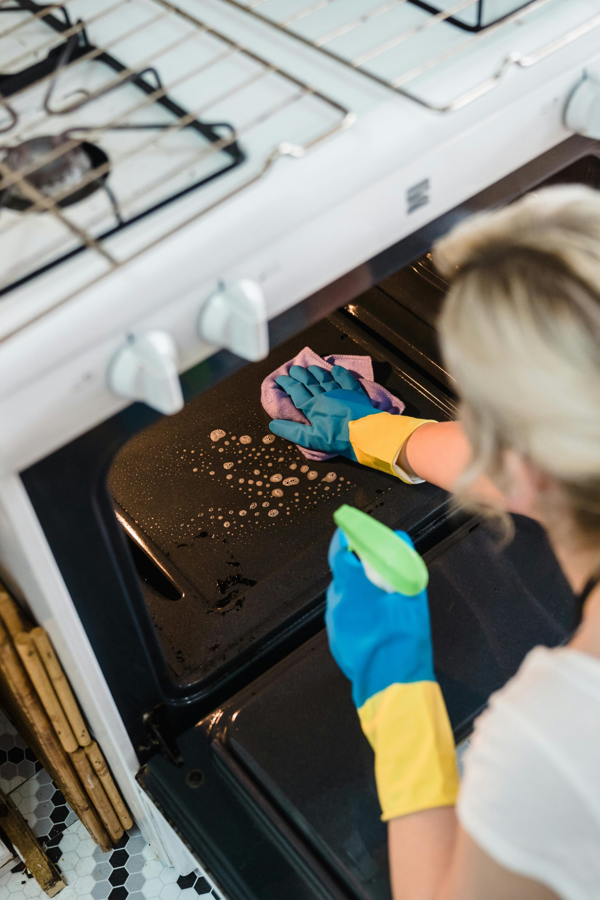 A woman wearing gloves cleans an oven in a domestic kitchen setting.